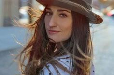 a woman with long hair wearing a fedora and looking at the camera while standing in front of a building