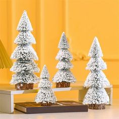 three snow covered trees sitting on top of a wooden table next to a lamp and book