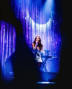 a woman standing in front of a stage with lights and curtains behind her, talking into a microphone