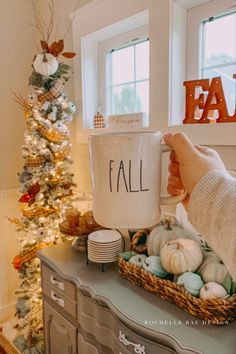 a person holding a coffee mug with the word fall on it in front of a christmas tree