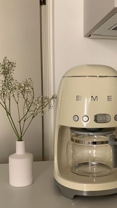 a cream colored coffee maker sitting on top of a counter next to a vase with flowers