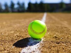 a green baseball sitting on top of a dirt field next to a white line and trees