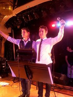 two men standing next to each other in front of microphones with arms raised up