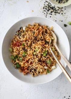 a white bowl filled with noodles and vegetables next to two silver spoons on a table