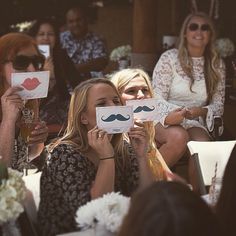 two women holding up signs with fake mustaches on them while others sit in the background