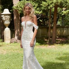 a woman in a white wedding dress standing on grass with trees and bushes behind her