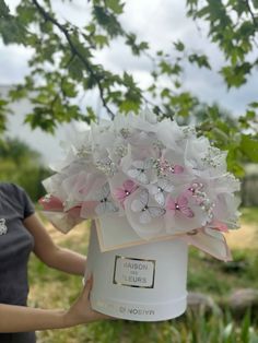 a woman is holding a white box with pink flowers in it while standing under a tree