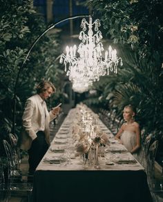 a man standing next to a woman at a long table in front of a chandelier