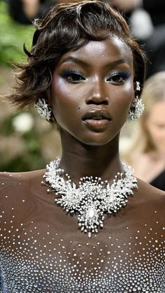 a model with dark skin and white makeup wearing an elaborate necklace and earrings on the runway