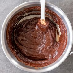 a metal bowl filled with chocolate frosting on top of a gray counter next to a spoon