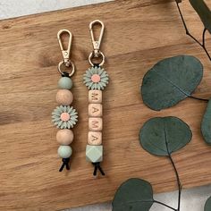 two keychains with flowers and letters on them sitting on a wooden table next to leaves