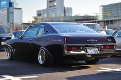 an old car is parked in a parking lot with other cars and buildings behind it