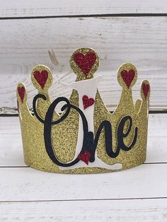 a gold glitter crown with hearts and the word one on it, sitting on a white wooden surface