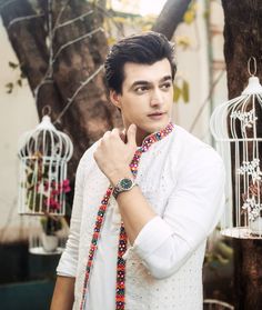 a man in white shirt and colorful beaded necklace next to bird cages with birds on them