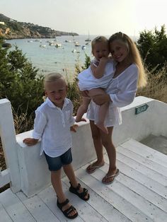 two girls and a boy are standing on a porch near the ocean with boats in the water