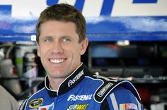 a close up of a person wearing a racing uniform and smiling at the camera with a nascar car in the background