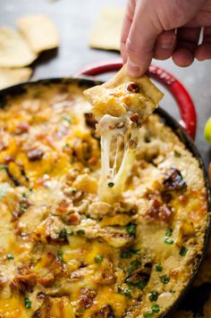 a hand holding a tortilla chip over a skillet filled with cheesy potatoes