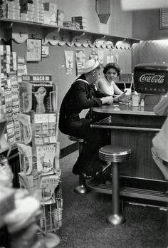 a man and woman sitting at a counter in a store with soda cans on the shelves