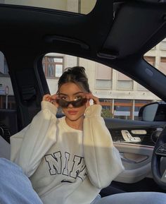 a woman sitting in the passenger seat of a car wearing sunglasses and a sweatshirt with letters on it