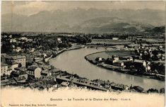 an old black and white photo of a river running through a town with mountains in the background