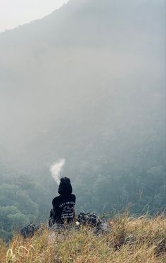 a person sitting on top of a hill next to a lush green hillside covered in fog