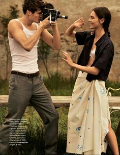 a man and woman standing next to each other in front of a wooden bench holding a camera