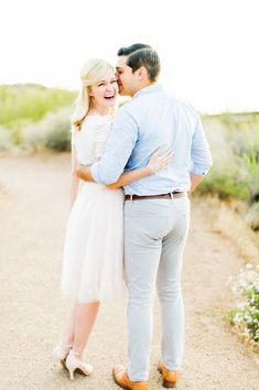 a man and woman standing together on a dirt road in front of some bushes with their arms around each other