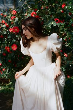 a woman wearing a white dress standing in front of red flowers with her hands on her hips