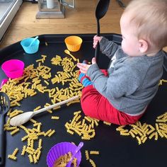 a baby sitting on top of a black tray filled with yellow pasta and measuring spoons