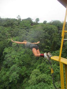 a woman hanging from the side of a zip line over a lush green forest filled with trees