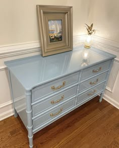 a blue dresser with gold hardware and a painting on the wall behind it in a corner