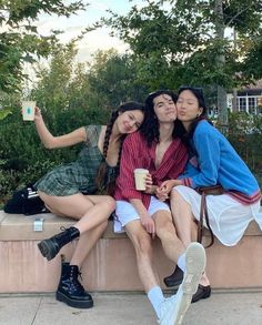 three young women sitting on a bench with one holding a coffee cup and the other taking a selfie