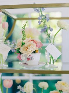 an arrangement of flowers in teacups on a glass shelf with cards and tags