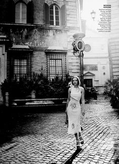 black and white photograph of woman walking on cobblestone street in front of building