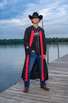 a man wearing a graduation robe and hat standing on a dock near the water with his hands in his pockets