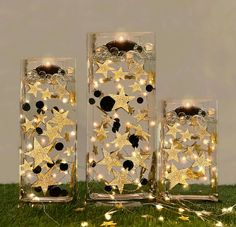 three clear glass vases with gold stars and lights in them on green grass next to wall