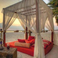 a bed sitting on top of a sandy beach next to the ocean with white drapes