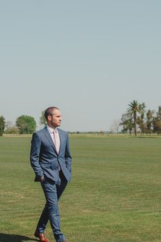 a man in a suit walking across a lush green field with palm trees behind him