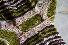 a knitted green and black striped sweater with pins in it's center, next to some knitting needles