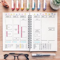 an open planner sitting on top of a wooden table next to pens and glasses,