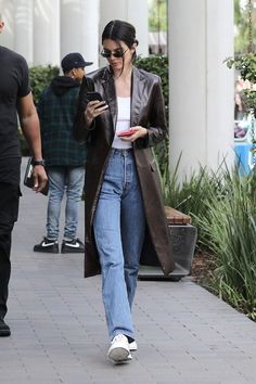 a woman walking down a sidewalk while looking at her cell phone and wearing jeans with white sneakers