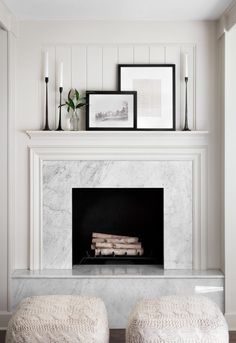 two white ottomans sit in front of a fireplace with pictures on the mantel above it