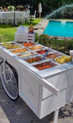 an outdoor buffet cart with food on it in front of a pool and lawn area