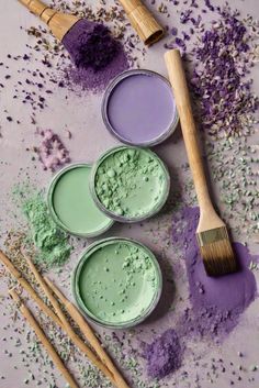 three paint cans with different colors and brushes next to them on a purple surface, surrounded by scattered lavender flowers