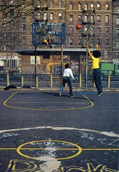 two young boys playing basketball on an outdoor court with graffiti written on the ground and buildings in the background