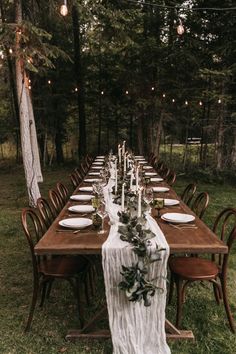 a long wooden table with white plates and greenery on it in the middle of a forest