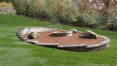 a fire pit surrounded by rocks and gravel in the middle of a grassy area with trees behind it