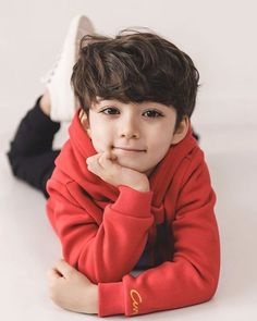 a young boy laying on the floor wearing a red sweater and black pants with his hand under his chin