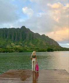 a woman standing on a dock next to a body of water with mountains in the background