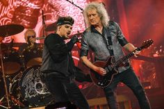 two men on stage with guitars in their hands and one holding an electric guitar up to his ear